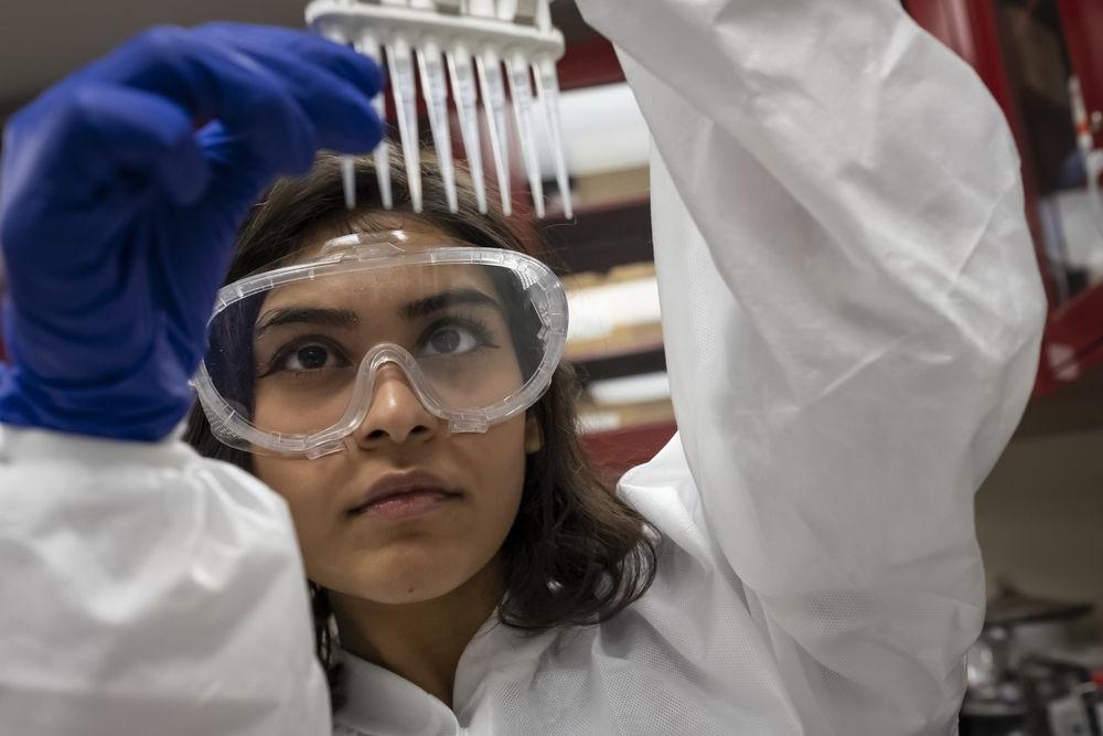 A student wearing a white lab coat and goggles inspects some test tubes.